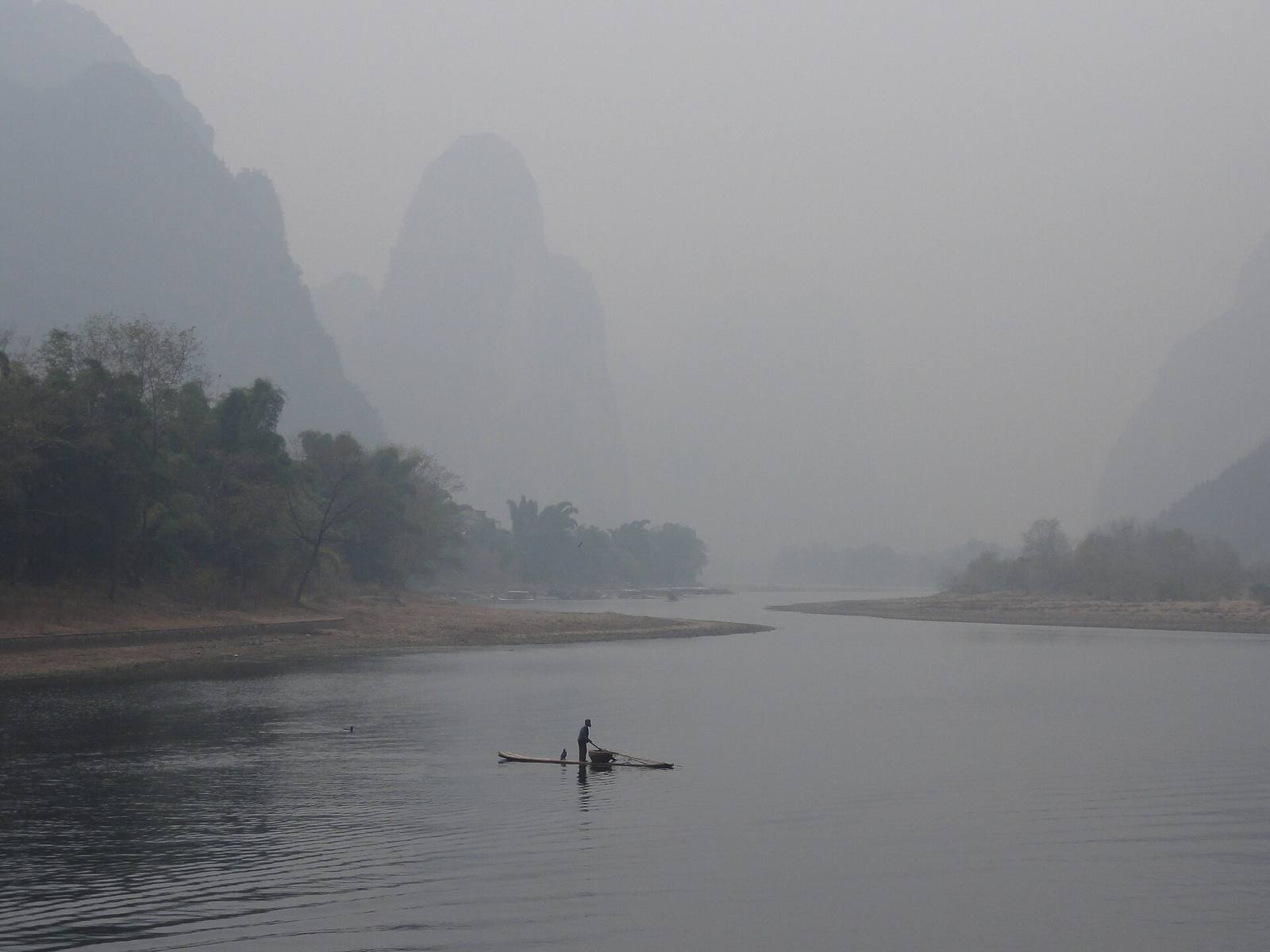 A lone cormorant fisherman poling a bamboo raft through dense morning mist on the Li River, karst peaks dissolving behind
