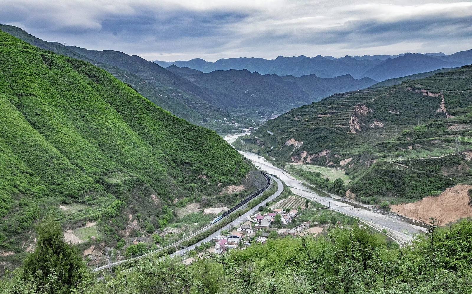 A long freight train winding through a deep green valley in the Qinling mountains